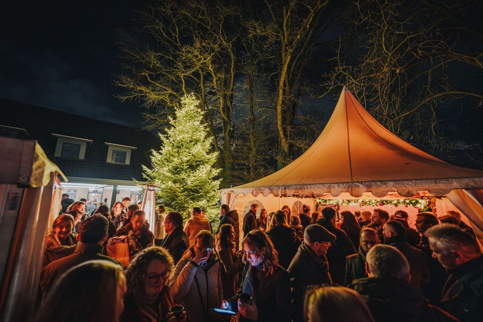 Gäste am Abend auf dem Weihnachtsmarkt am Patentkrug, mit beleuchtetem Tannenbaum und Zelt